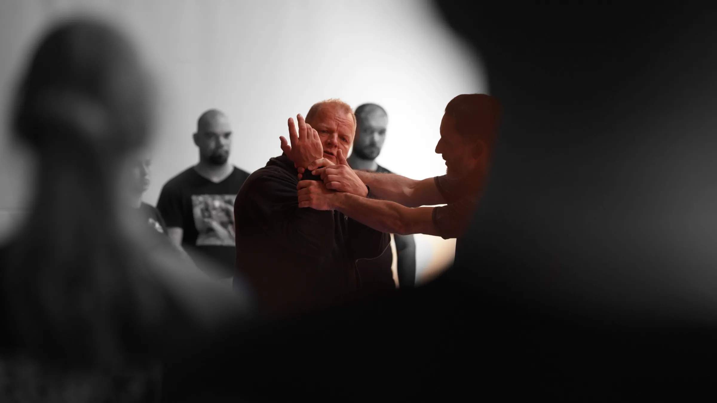 A senior instructor demonstrating a technique hand to hand while students watch, backlit in a plain hall
